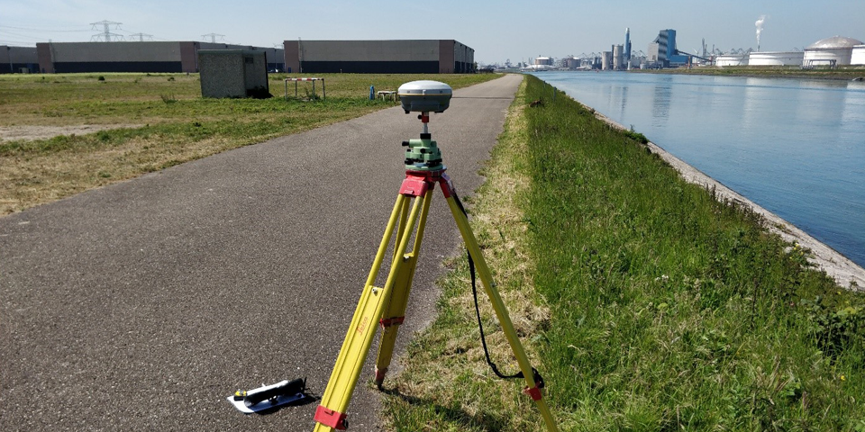 Landmeetkundige apparatuur op een weg naast een waterkant met een stedelijk skyline op de achtergrond.
