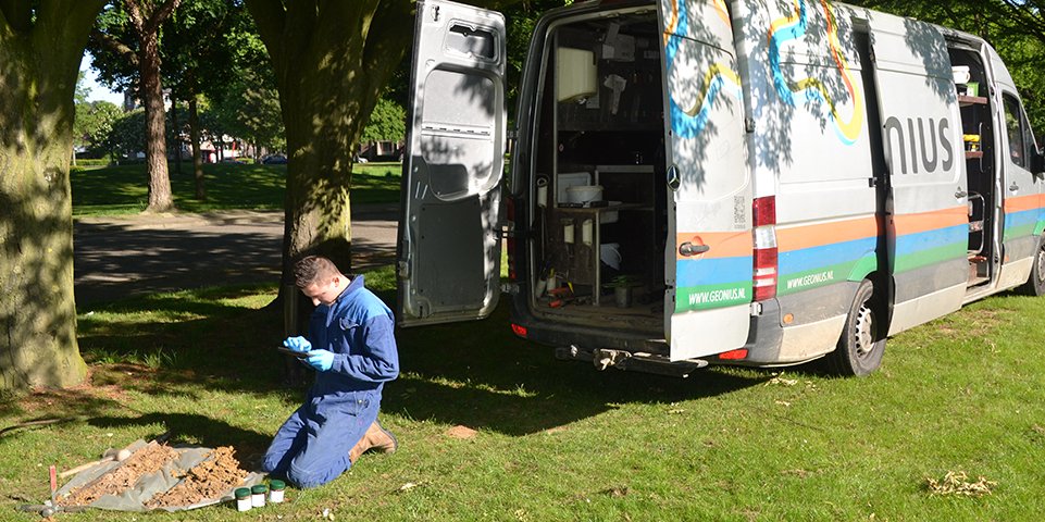 Een onderzoeker in een blauw overall knielt op het gras bij een boom, met een tablet in de hand, naast een gehuurde onderzoeksbus.