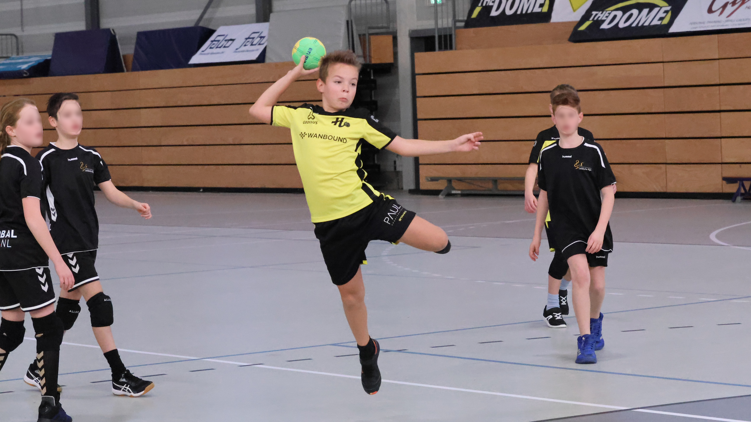 Kinderen spelen handbal in een sporthal, met een jongen die springt en de bal vasthoudt.