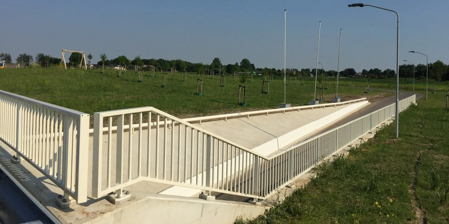 Modderbad met witte railing en straatverlichting, omliggende groene grasvelden en jonge bomen, in een open veld onder een blauwe hemel.