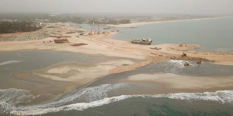 Larende kustlijn onder constructie, met schepen en zware machines op de zandbodem en vlakbij de zee.