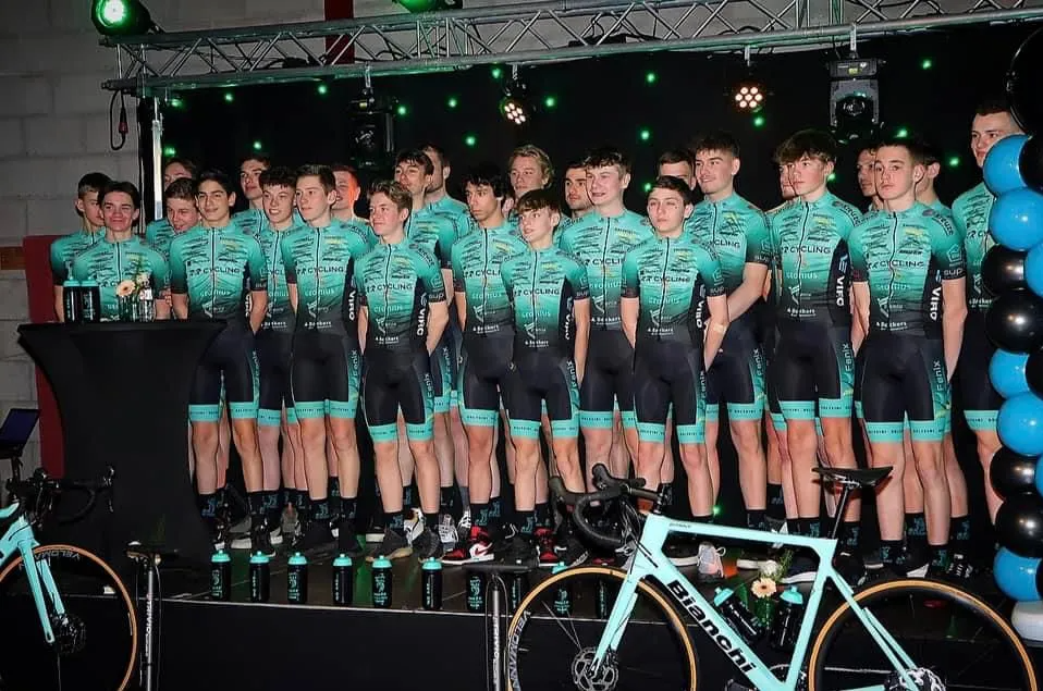 Een groep jonge wielrenners in uniform poseert op een podium, met fietsen en drankflessen voor hen, onder groene en gekleurde lichten tijdens een feest of teamviering.