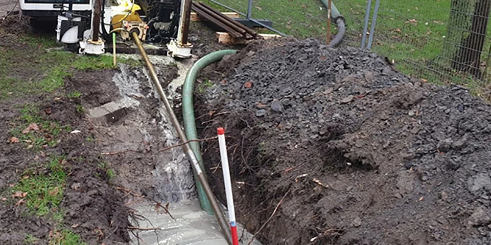 Ondergrondse installatie in de bouwput met buizen en graafwerk, omgeven door aarde en een hek.