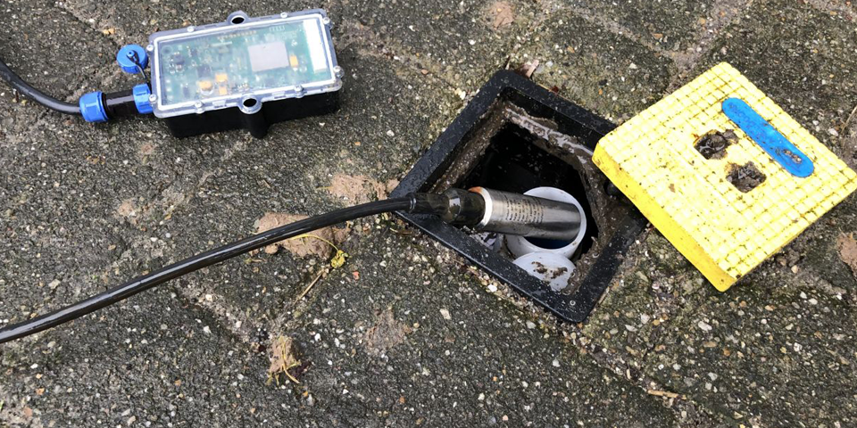 Een open waterleidingput met een ontstoppingsapparaat en een elektronische meter buiten op een weg of stoep, omringd door zand en kleine stenen.