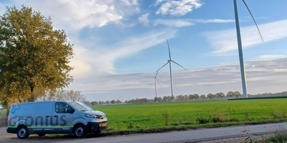 Een groen open veld met windmolens op de achtergrond, een blauwe bestelbus geparkeerd langs de weg en een grote boom aan de linkerkant.