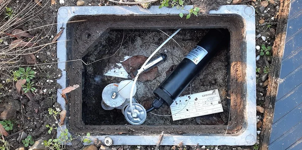 Een open betonput met daarin elektrische componenten, draden, een waterdichte lamp en een co2 sensor in een tuin of buitenruimte.
