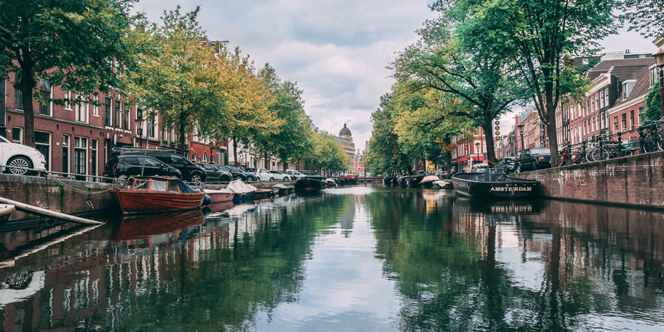 Gracht in Amsterdam met boten, ouderwetse gebouwen, bomen en een brug in de verte.
