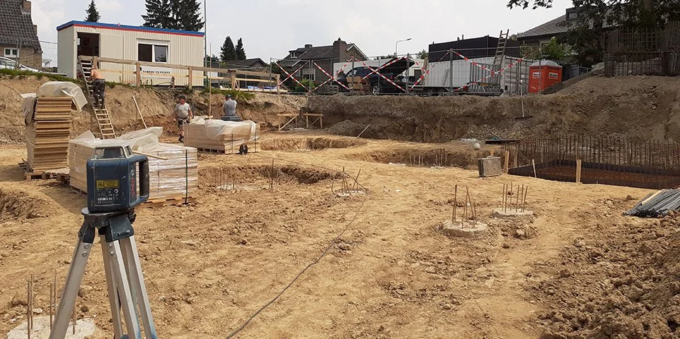 Een bouwplaats met ondergrondse funderingswerkzaamheden, bouwmaterialen en werkers. Er is een landmeter met een laser op een statief in de voorgrond.