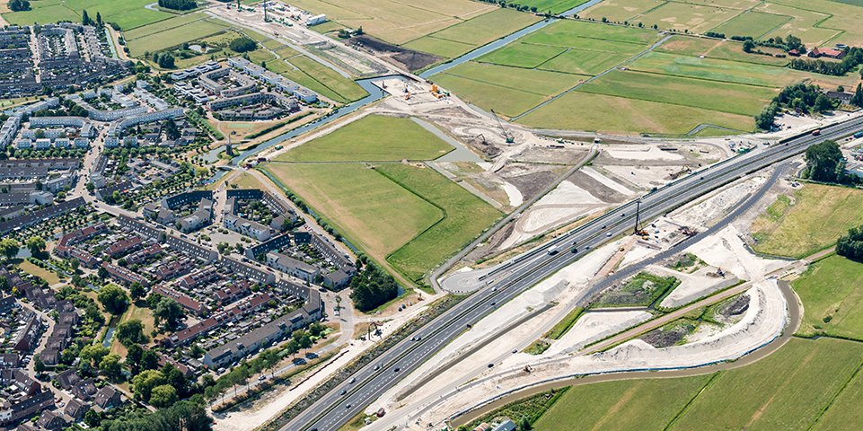 Luchtfoto van een snelweg die onder constructie is, met bouwmachines en wegenwerken, vlakbij een woonwijk en groene velden.