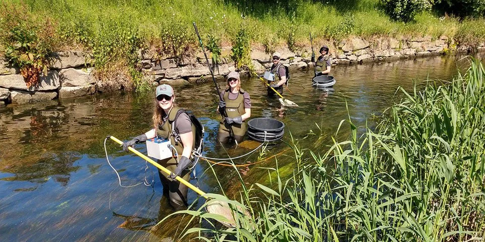 Vier mensen in een waterweg met wetenschappelijke uitrusting, met netten en meetapparatuur.