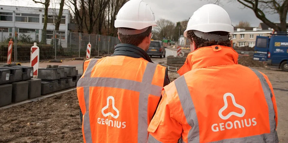 Twee bouwvakkers in oranje veiligheidsjassen en helmen staan op een bouwplaats.