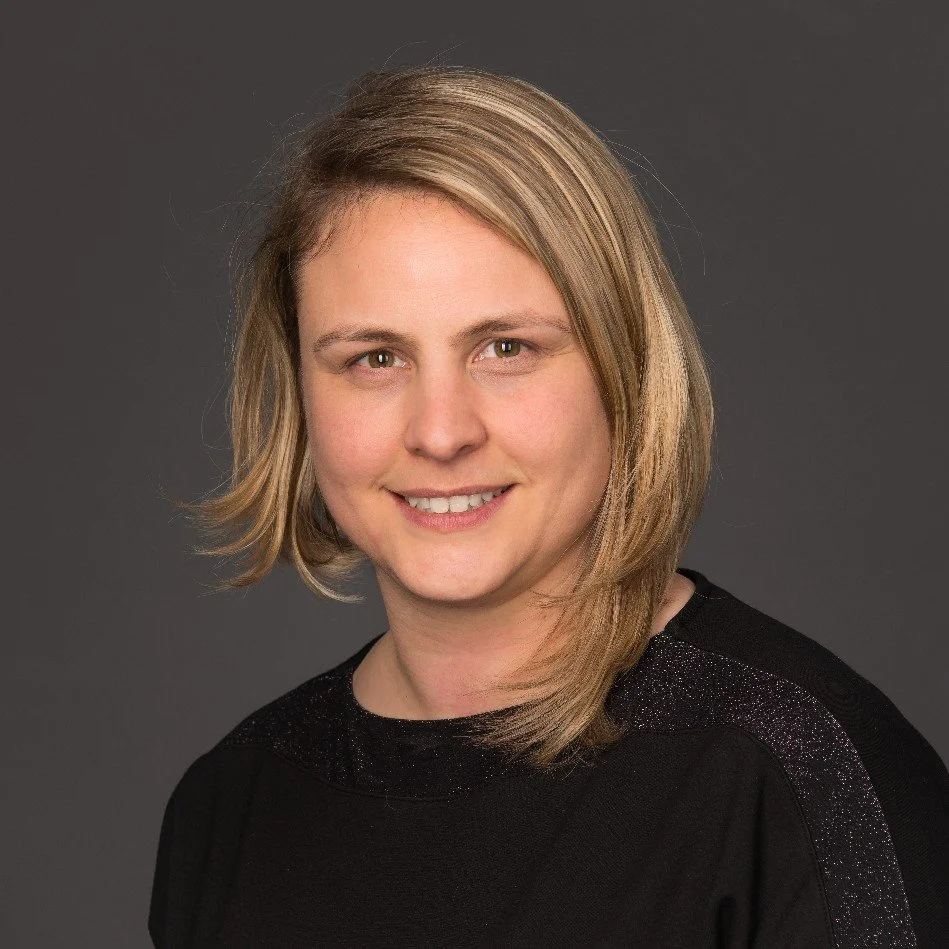 Portret van een vrouw met blond, halflange haar, gekleed in een zwarte top, glimlachend voor een grijze achtergrond.
