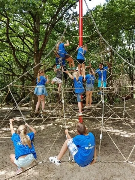 Jeugdgroep die op een touwen klimgordel oefenen in een bos, iedereen draagt blauwe shirts.