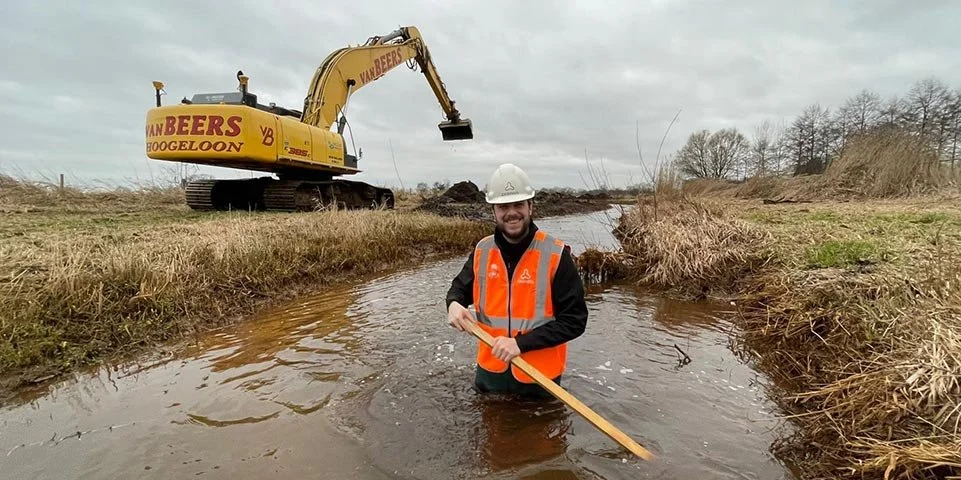 Man met een veiligheidshelm en een oranje veiligheidsvest staat in een smalle waterweg, een graafmachine op de achtergrond.