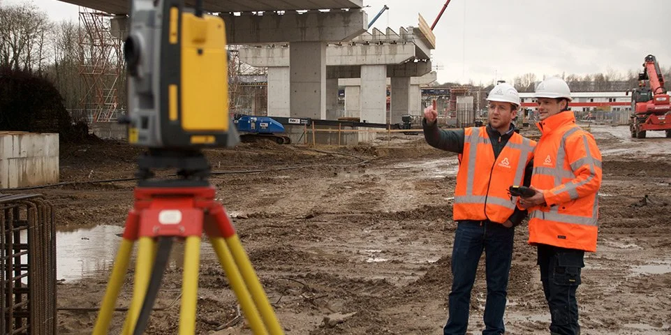 Twee bouwvakkers in oranje veiligheidsjassen en helmen die op een bouwplaats staan, terwijl een van hen wijst naar iets in de verte.