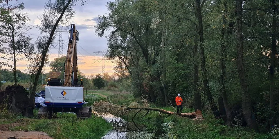 Een graafmachine aan de oever van een rivier met een man in een oranje jas en witte helm in een bosrijke omgeving.