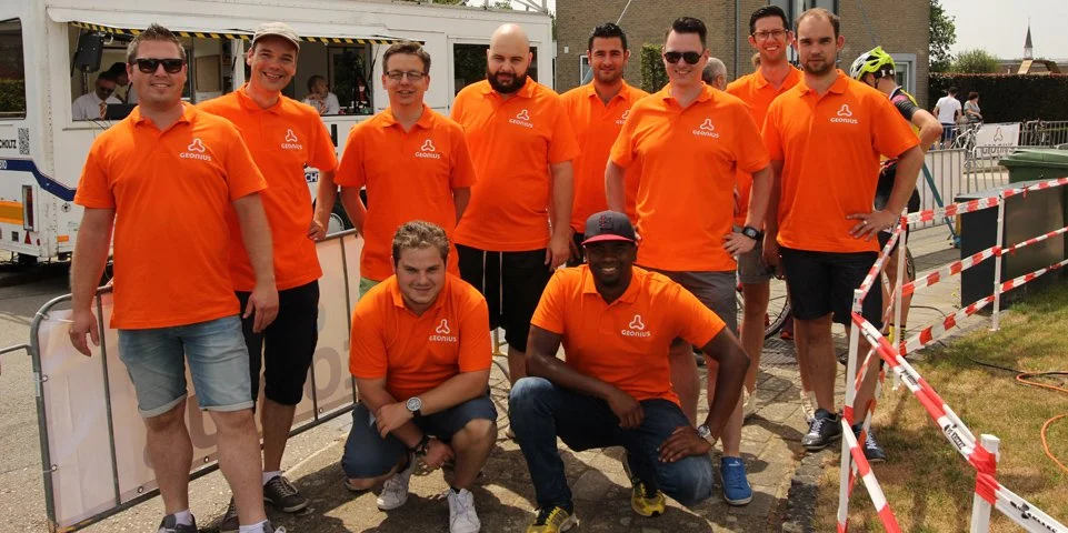 Tien mannen en een jongen in oranje GEOUS shirts verzamelen zich buiten tijdens een evenement, enkele mensen werken op de achtergrond.