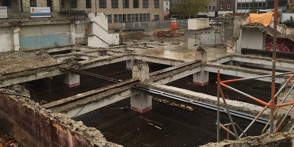 Constructieplaats met verweerde betonvloeren en ondersteuningen, bouwplanken en steigers zichtbaar.