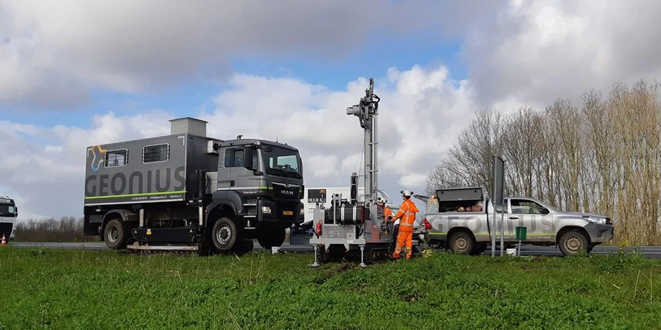 Werkers in oranje veiligheidskleding werken aan meetapparatuur naast een geleidewagen op een weg, met bomen en een partly cloudy hemel op de achtergrond.