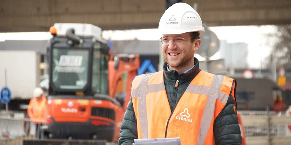Een man in een veiligheidsvest en helm op een bouwplaats, lachend.