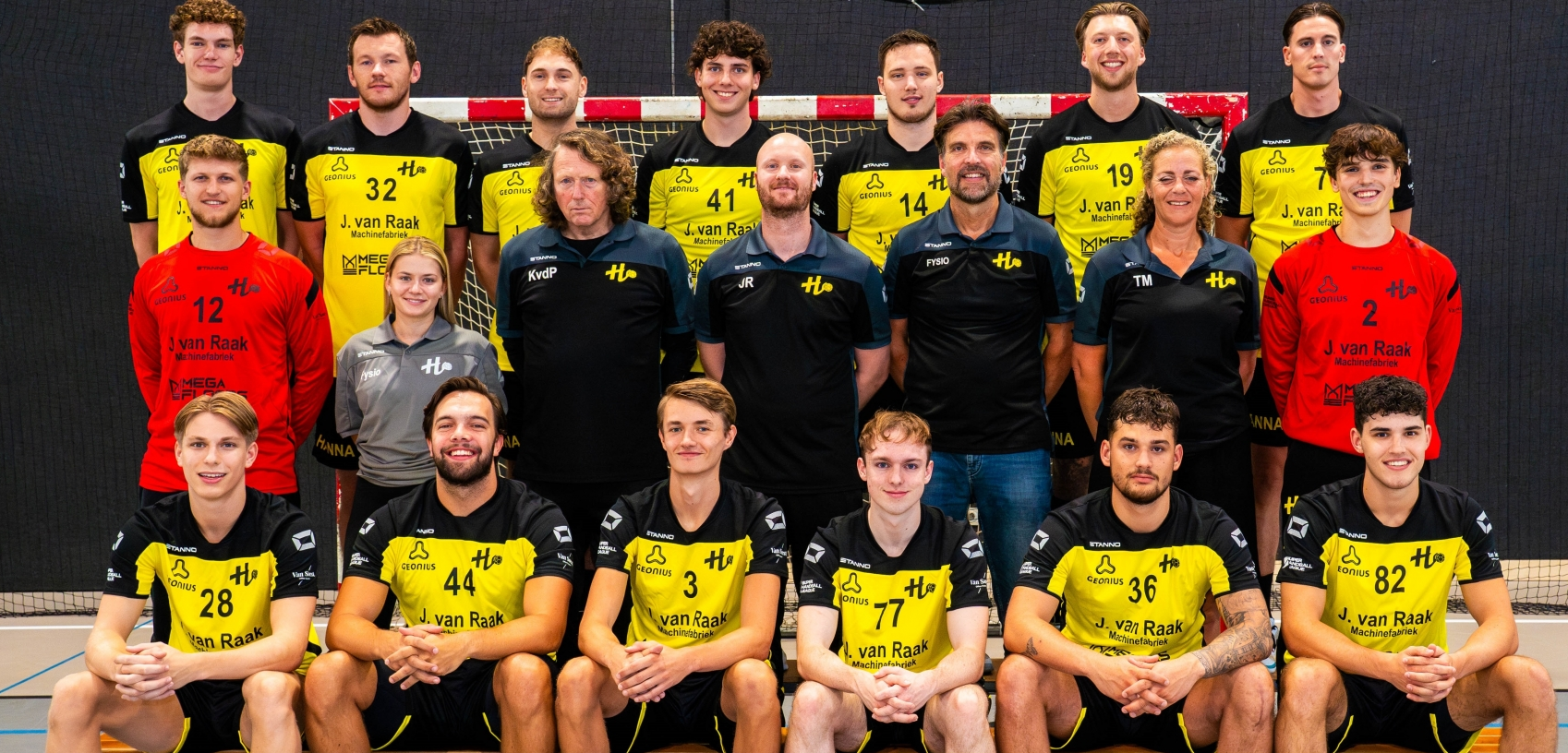 Een team van mannen en vrouwen dat handbal speelt, gekleed in gele en zwarte jerseys, poseert voor een groepsfoto in een sporthal met een handbaldoel op de achtergrond.