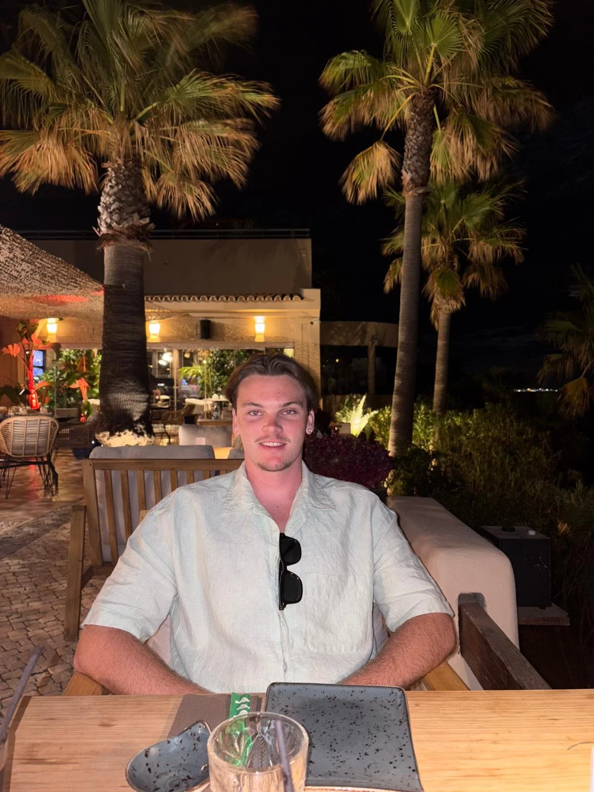 A young man sitting at a restaurant table outdoors at night, with palm trees and warm lighting in the background.