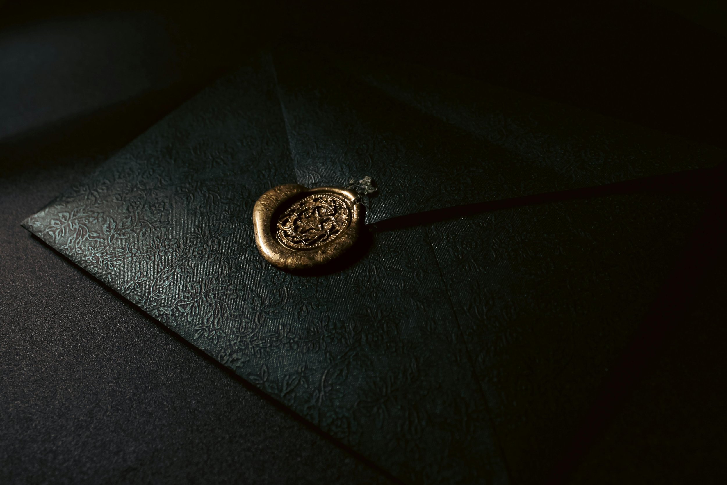 Ein schwarzer Briefumschlag mit einem goldenen Siegel und eingraviertem Muster, liegt auf einer dunklen Oberfläche.