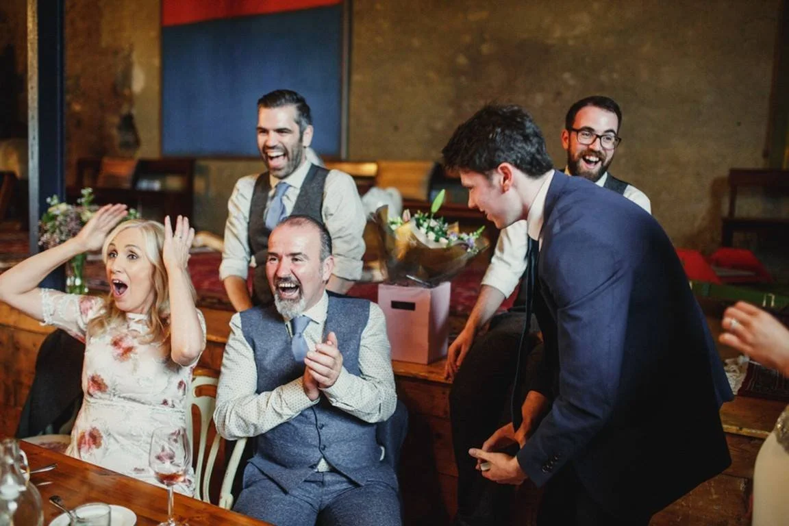 Group of five adults joyfully celebrating, with two seated at a table and three standing behind them, in what appears to be a lively party or wedding reception.