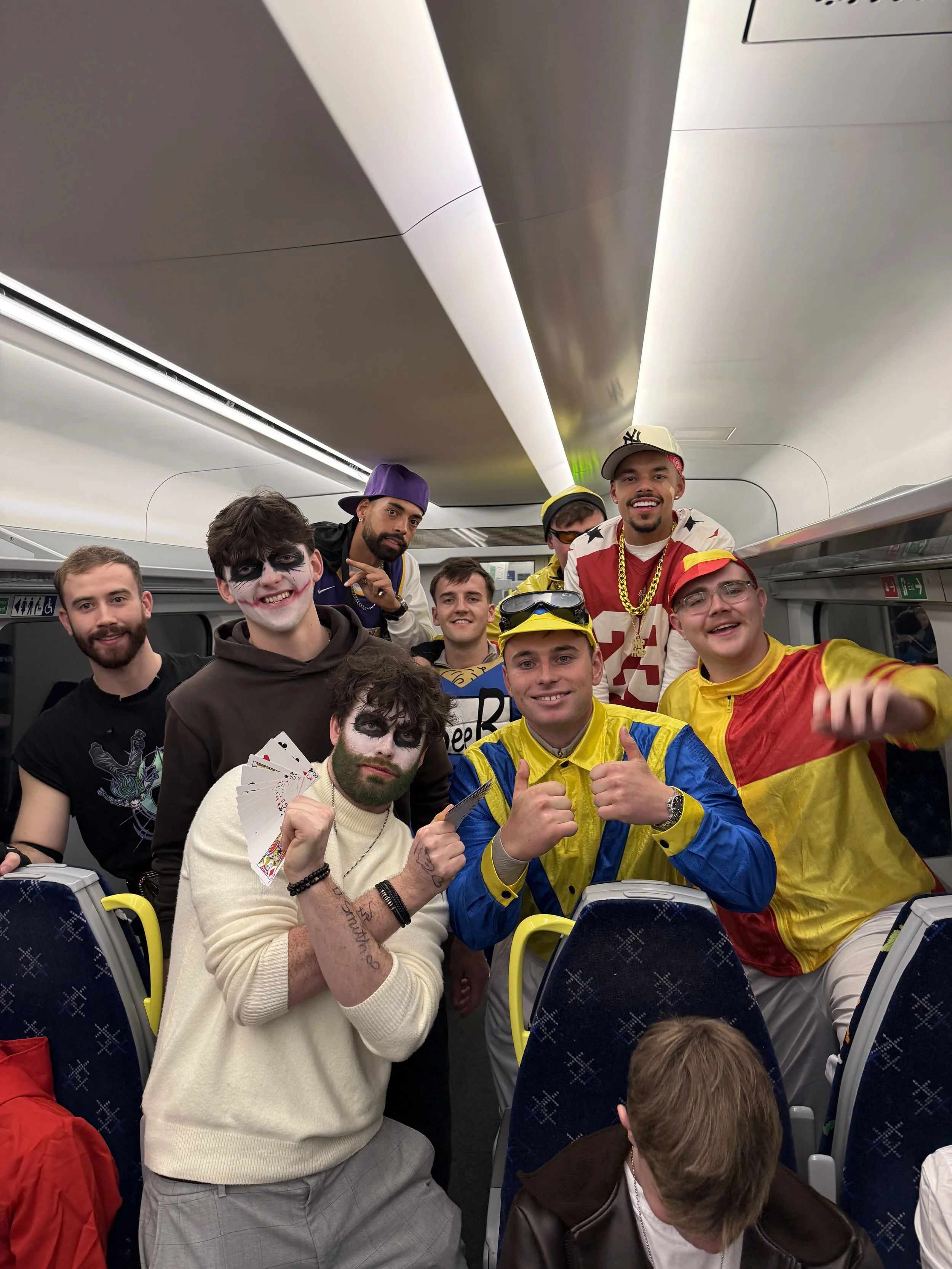 A group of young men, some in costume and others in casual clothes, are posing for a photo inside an airplane, some smiling and making gestures like thumbs up.
