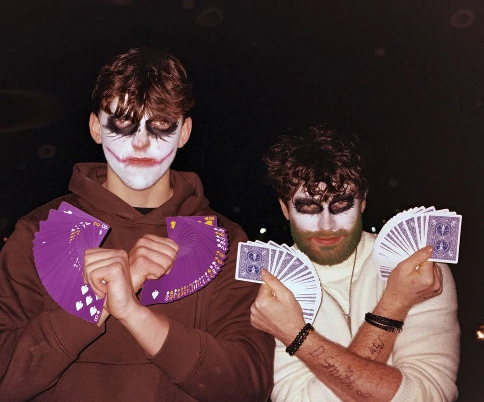 Two young men with face paint resembling Joker characters from Batman, holding fan of purple playing cards. They are dressed casually, with one in a brown hoodie and the other in a white sweater, against a dark background.