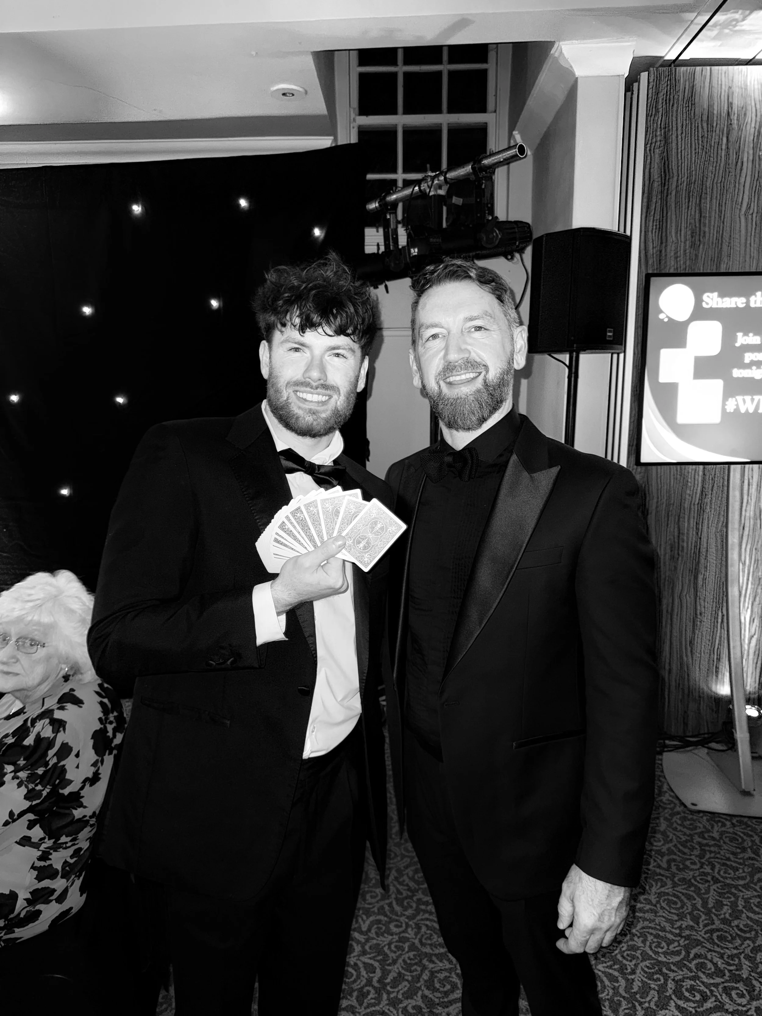 Two men in tuxedos smiling at the camera; one holding playing cards. They are at an indoor event with a black backdrop decorated with small lights.