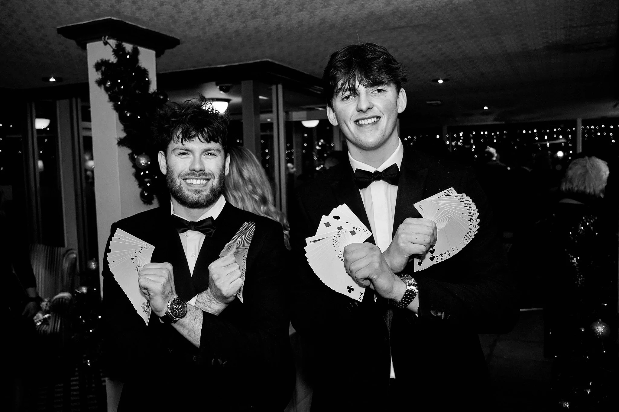 Two young men in tuxedos holding playing cards and smiling at a party with holiday decorations in the background.