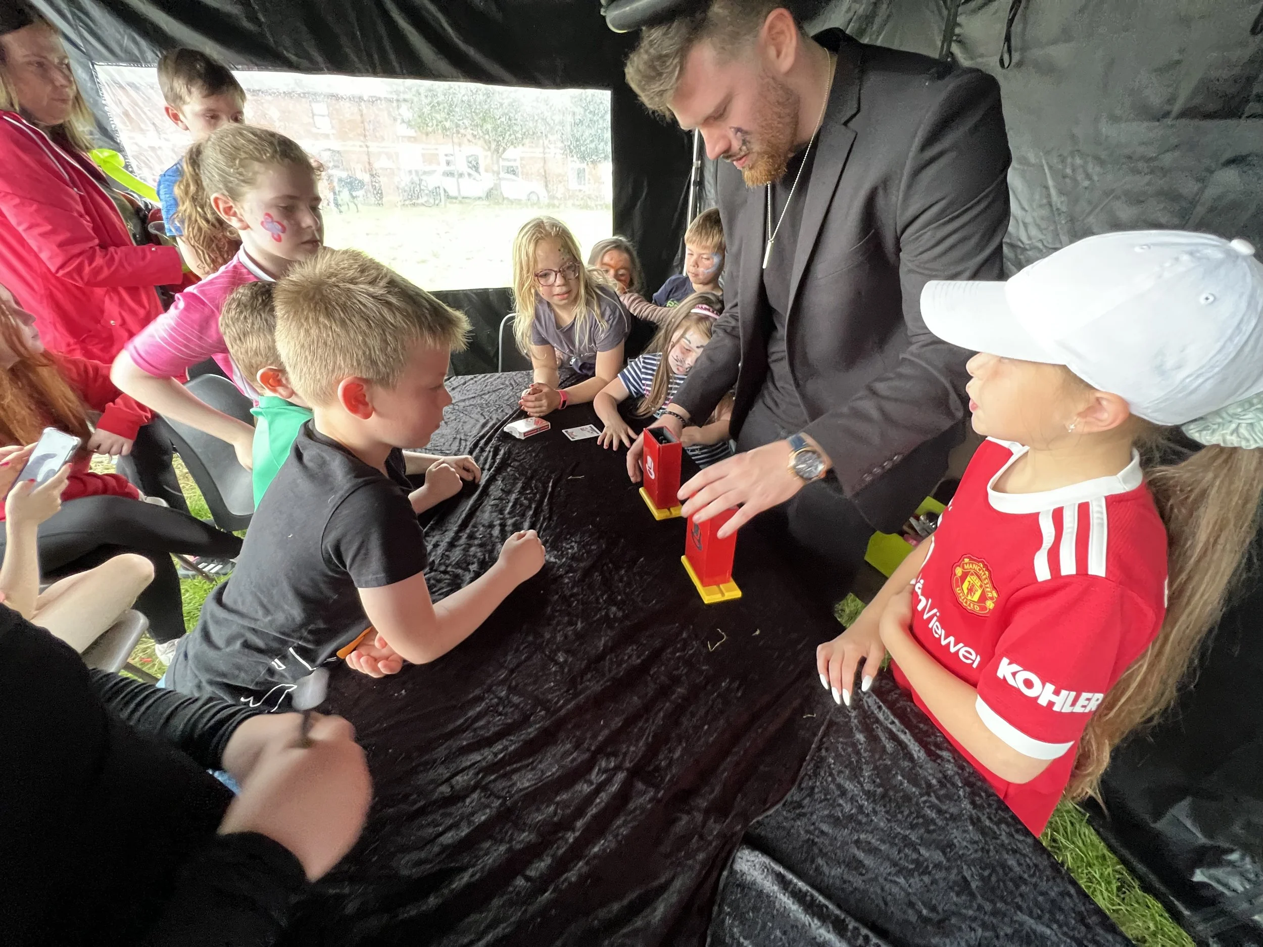 Children gathered around a table watching a man in a black suit perform a magic trick with two red boxes during an outdoor event under a black tent.