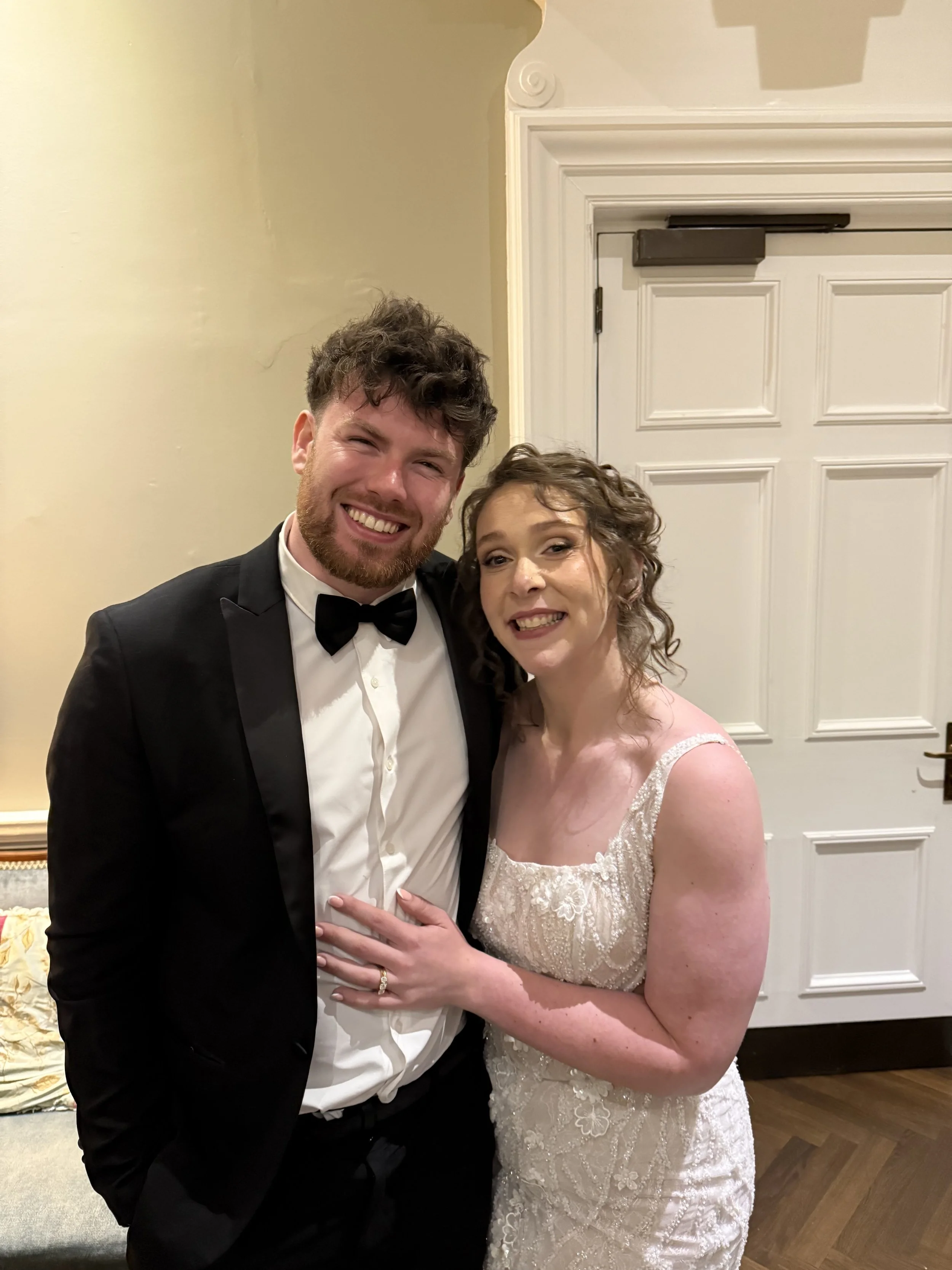 A smiling couple dressed in formal wedding attire, the man in a black tuxedo with a bow tie, and the woman in an elegant white lace wedding gown, standing in front of a white door.