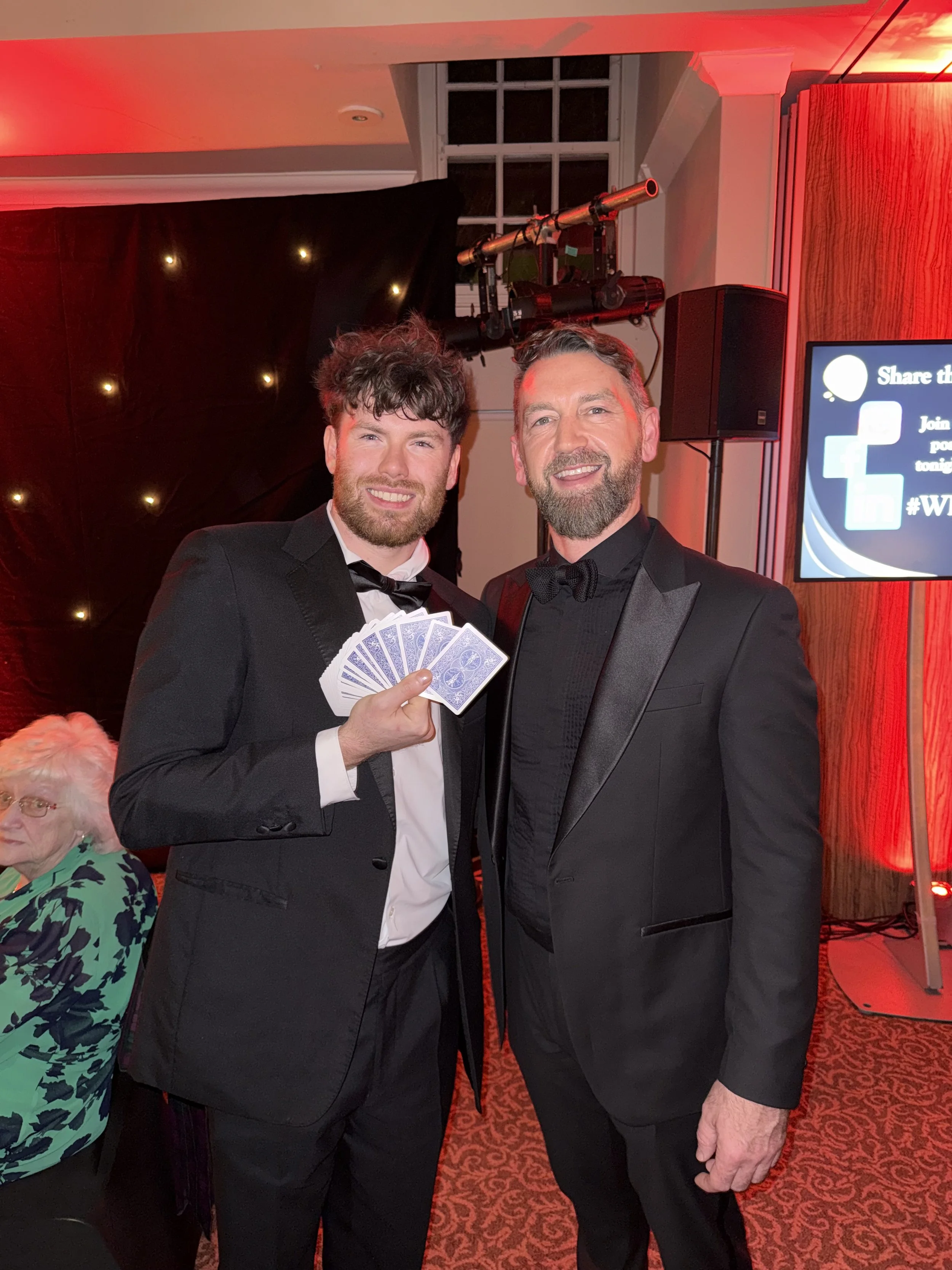 Two men in tuxedos, one holding playing cards, pose for a photo at a formal event with red lighting and a decorated background.