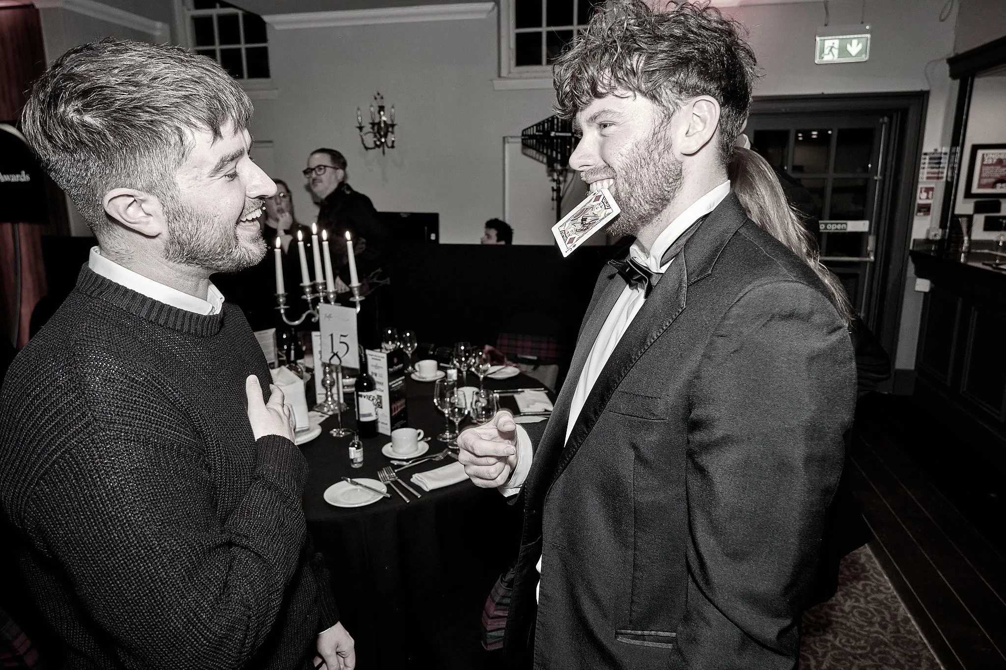 Two men in formal attire laughing and engaging in conversation at a social event. One has a card in his mouth, and the other has his hand on his chest, suggesting a moment of amusement or surprise.