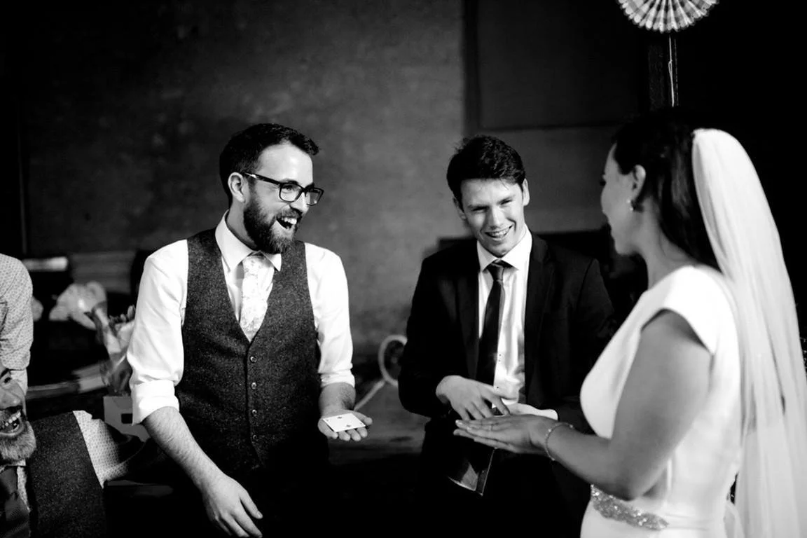 People smiling and engaging in conversation at a wedding reception, a woman in a wedding dress and veil talking to two men, one in a vest and glasses and the other in a suit.