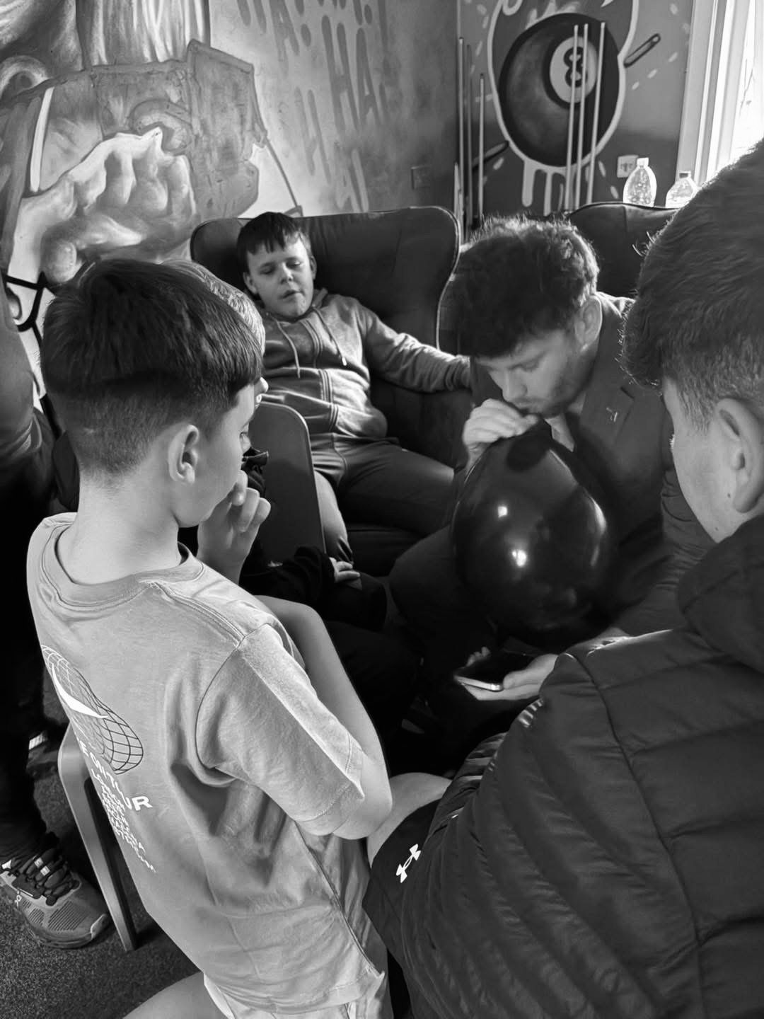 A group of young boys and a few men gathered closely together in a room, some sitting and some standing. One boy is seated with his hand near his mouth, while the others appear to be focused on something in the center of the group, which involves a balloon. The room has a mural on the wall and a couple of water bottles on a table in the background.