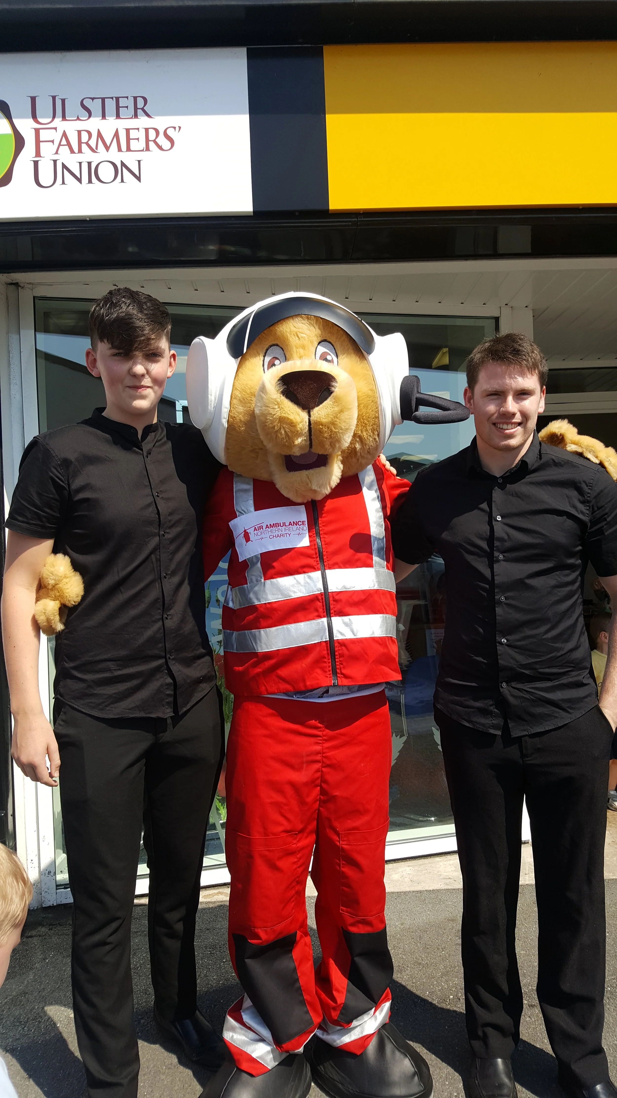 Two young men and a lion mascot in a red and white uniform standing outside a building with a sign for Ulster Farmers Union.