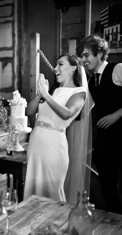 A woman in a wedding dress and a man in formal attire celebrating at a wedding reception, with a wedding cake on the table.
