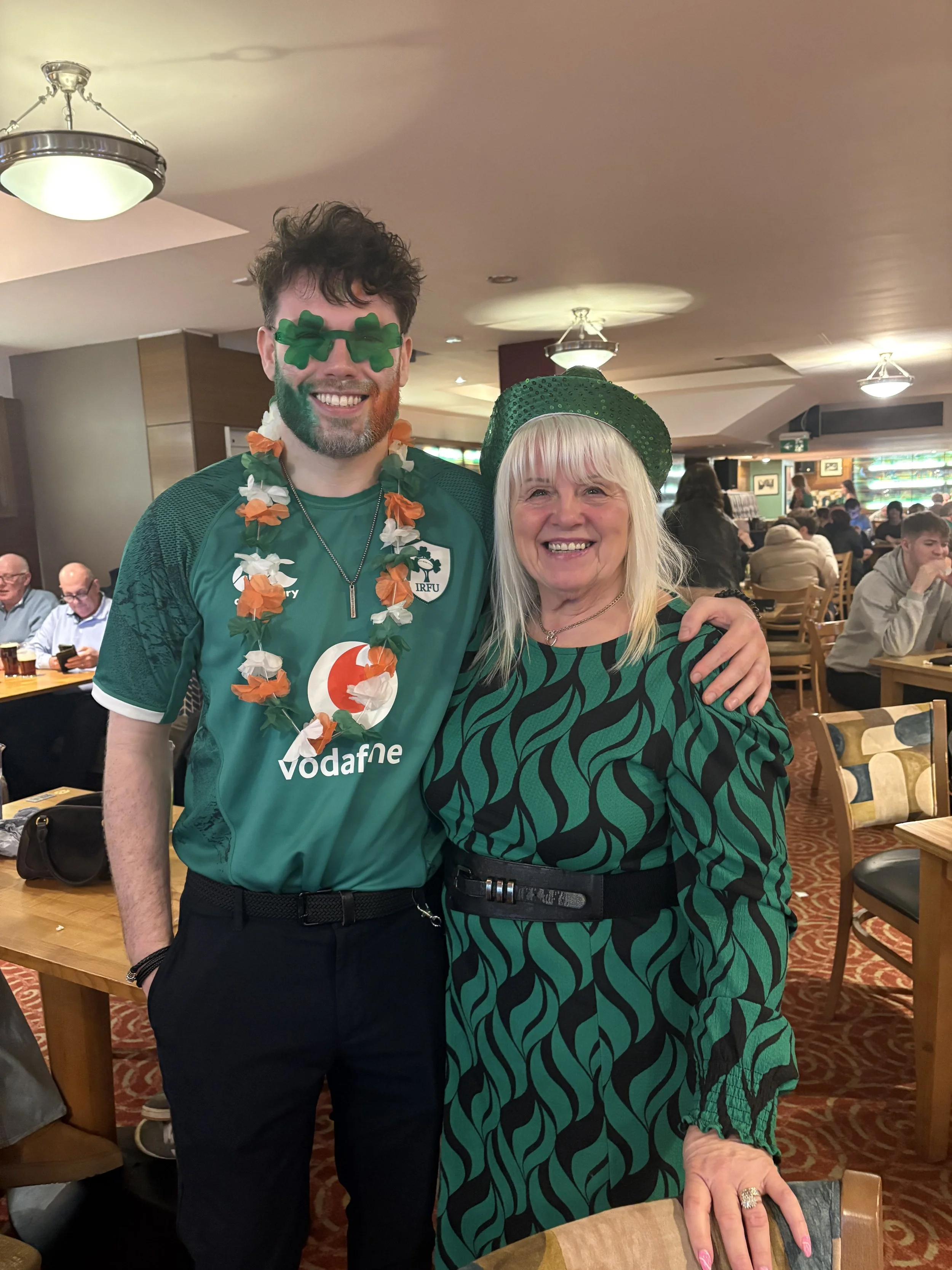 Two people, a man and a woman, celebrating St. Patrick's Day at a restaurant. The man is wearing shamrock-shaped sunglasses, a green Irish rugby jersey, and a floral lei. The woman is wearing a green and black patterned dress, a black belt, and a green hat. They are smiling, standing close together, with the woman resting her hand on a chair.