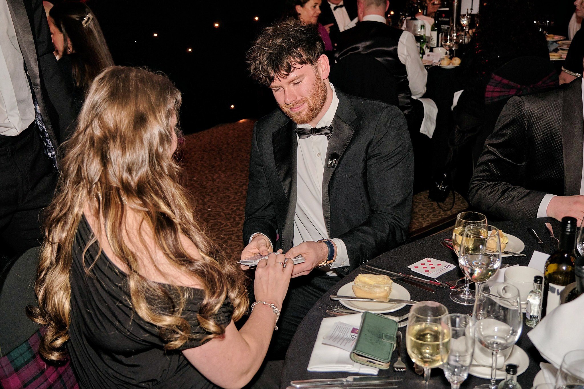 A man in a tuxedo with a bow tie is sitting at a table, looking at a woman who is showing him something on her phone, in a formal dinner setting.