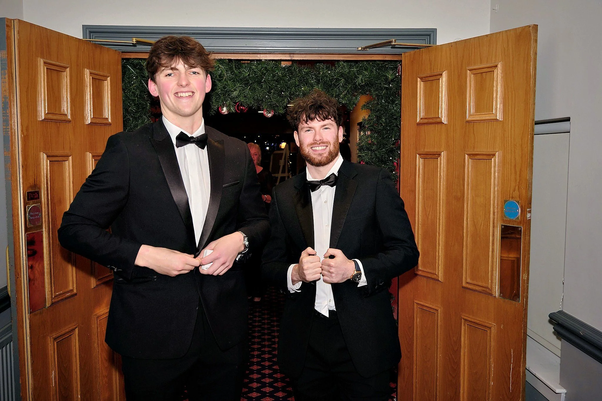 Two young men in tuxedos smiling at the camera, standing in front of a festive doorway decorated with green foliage and red ornaments.