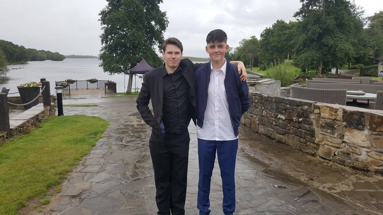 Two young men standing outdoors by a lake, with trees and a stone walkway, wearing semi-formal attire.