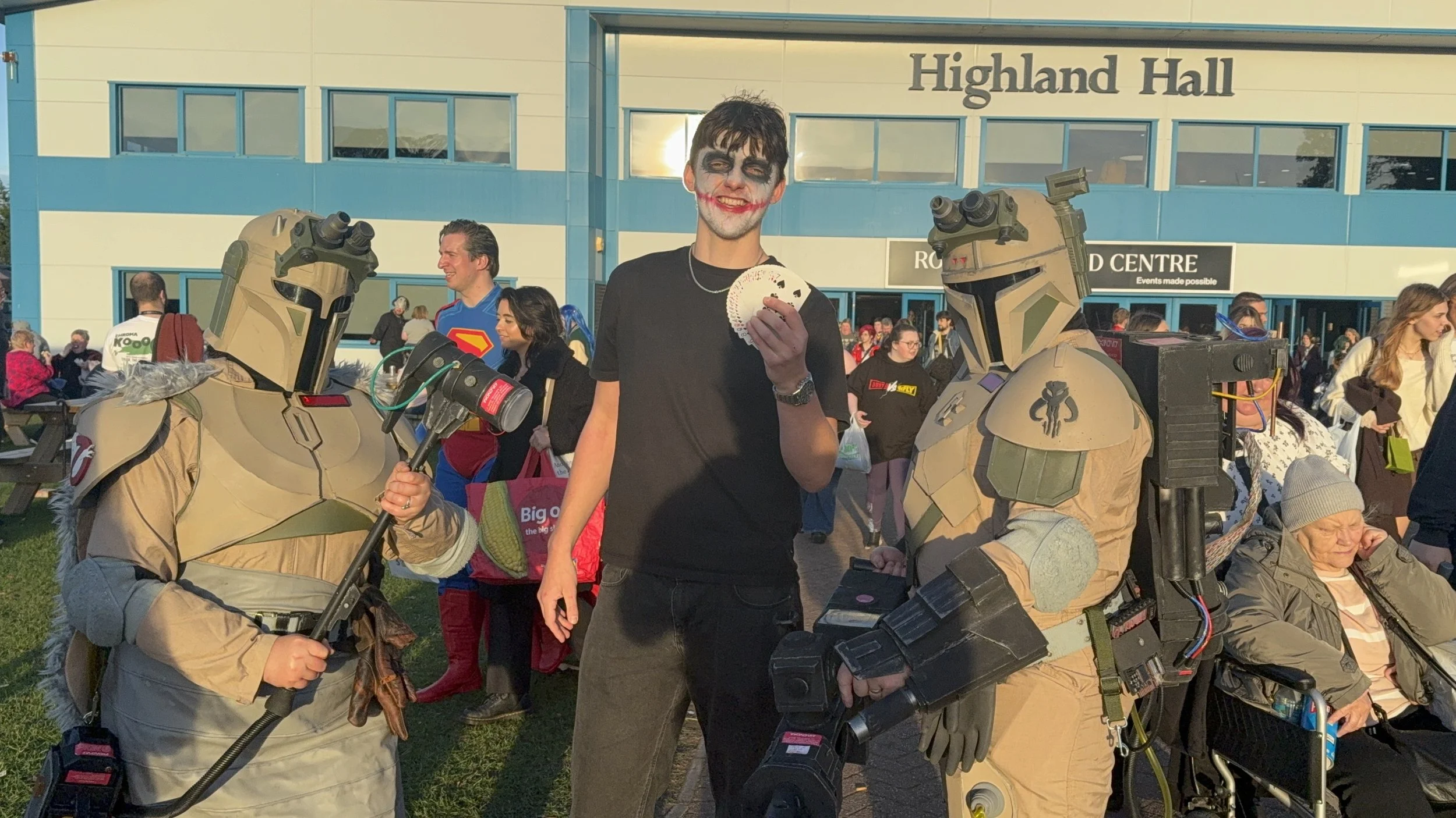 A young man dressed as the Joker with face paint, holding playing cards, standing among people and costumed individuals in military armor at an outdoor event with Highland Hall in the background.