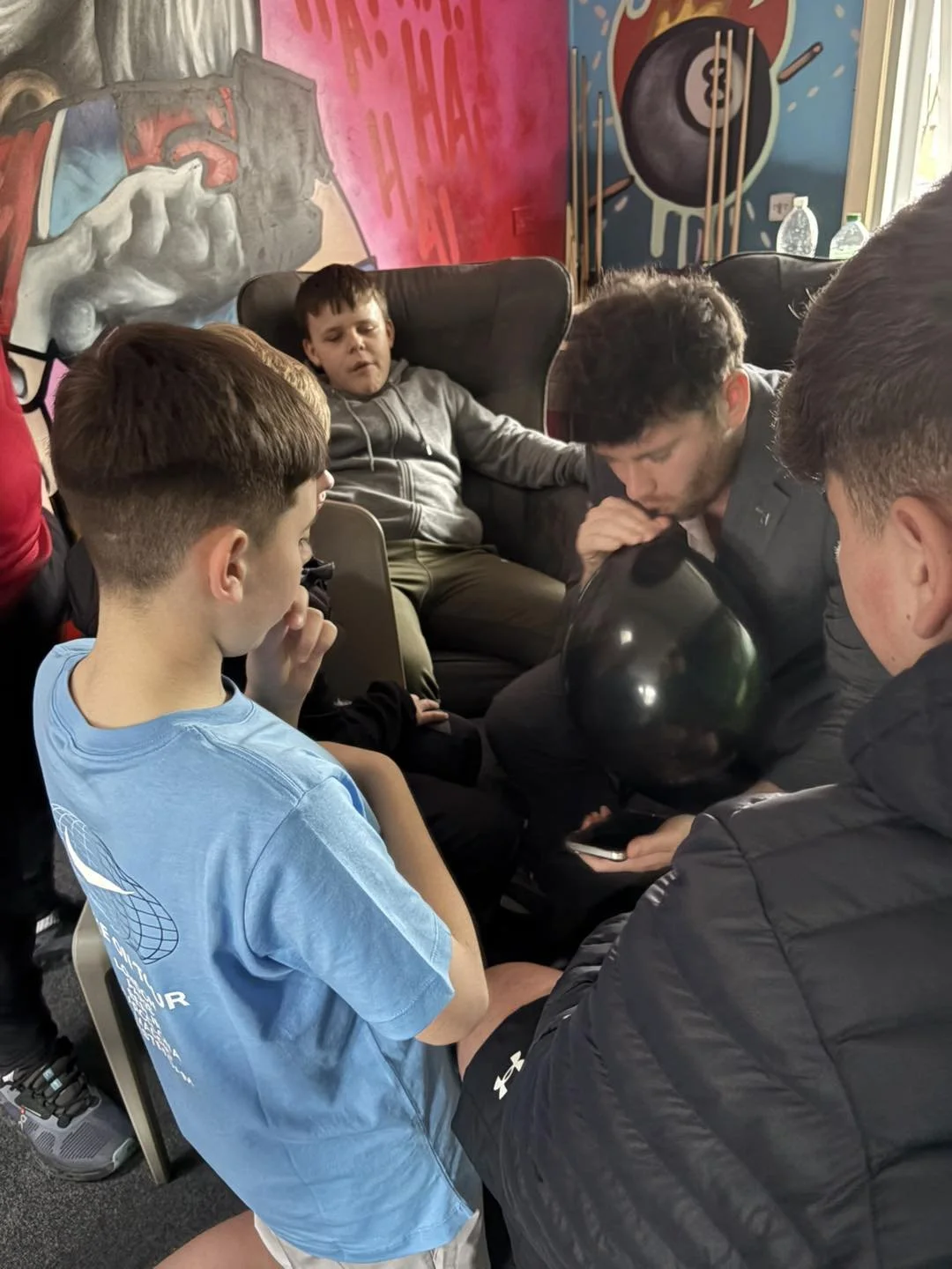Group of boys and young men gathered around in a room with graffiti arts on the walls, one boy in a gray hoodie sitting relaxed in a chair, and others focused on a smartphone.
