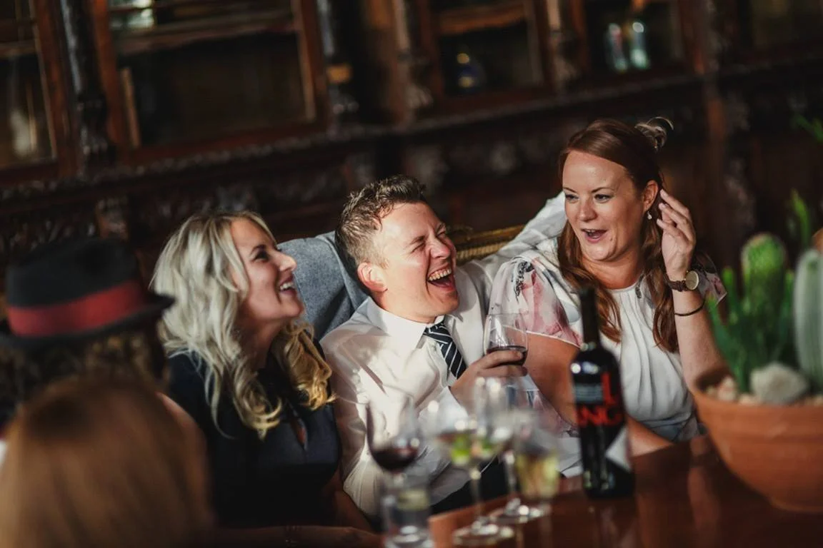 Four people sitting close together at a dinner table, laughing and chatting, with drinks and a bowl of flowers in the foreground, in a cozy, wood-paneled setting.