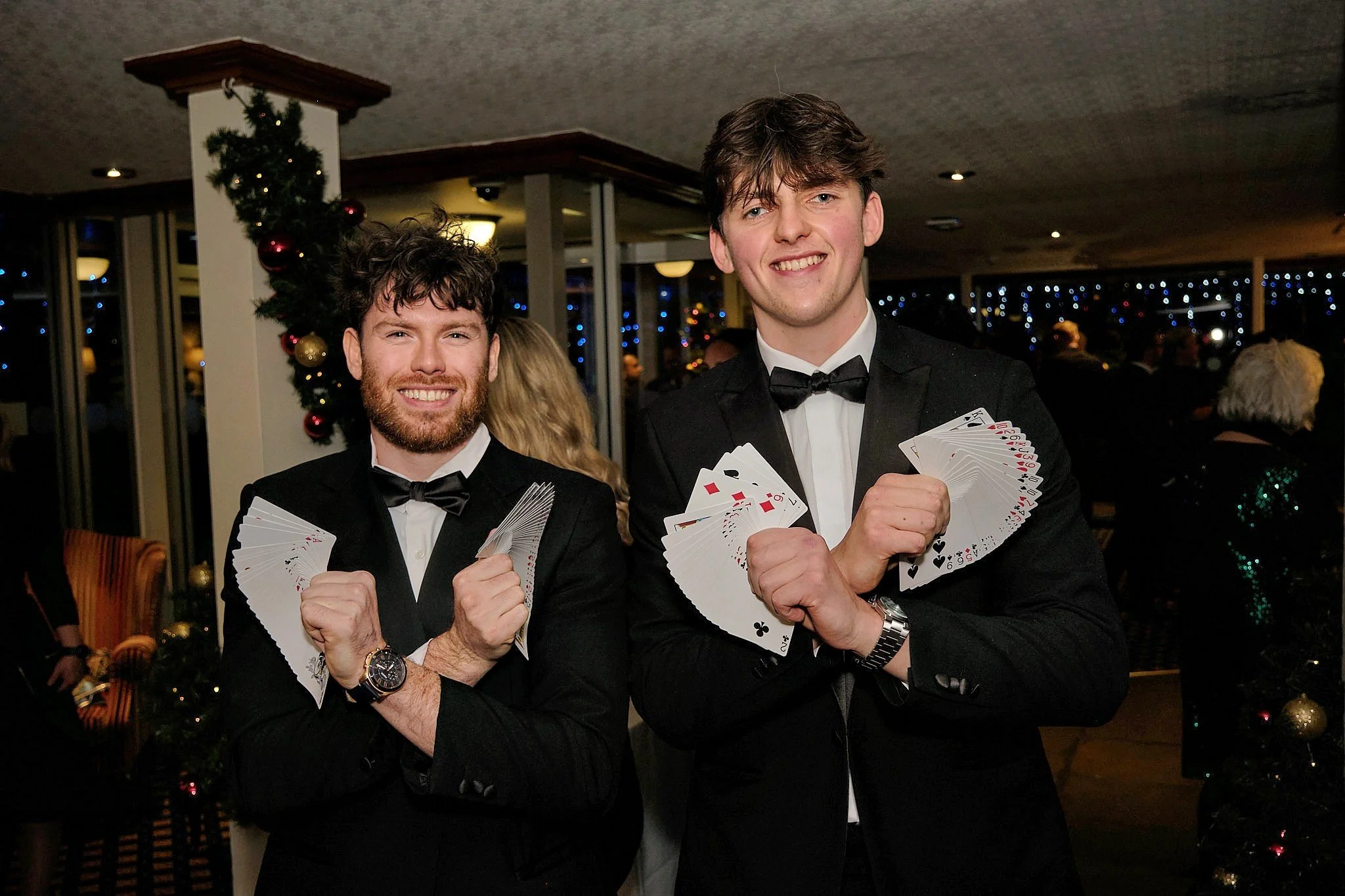 Two men dressed in tuxedos holding fanned playing cards at a festive event with Christmas decorations and other people in the background.