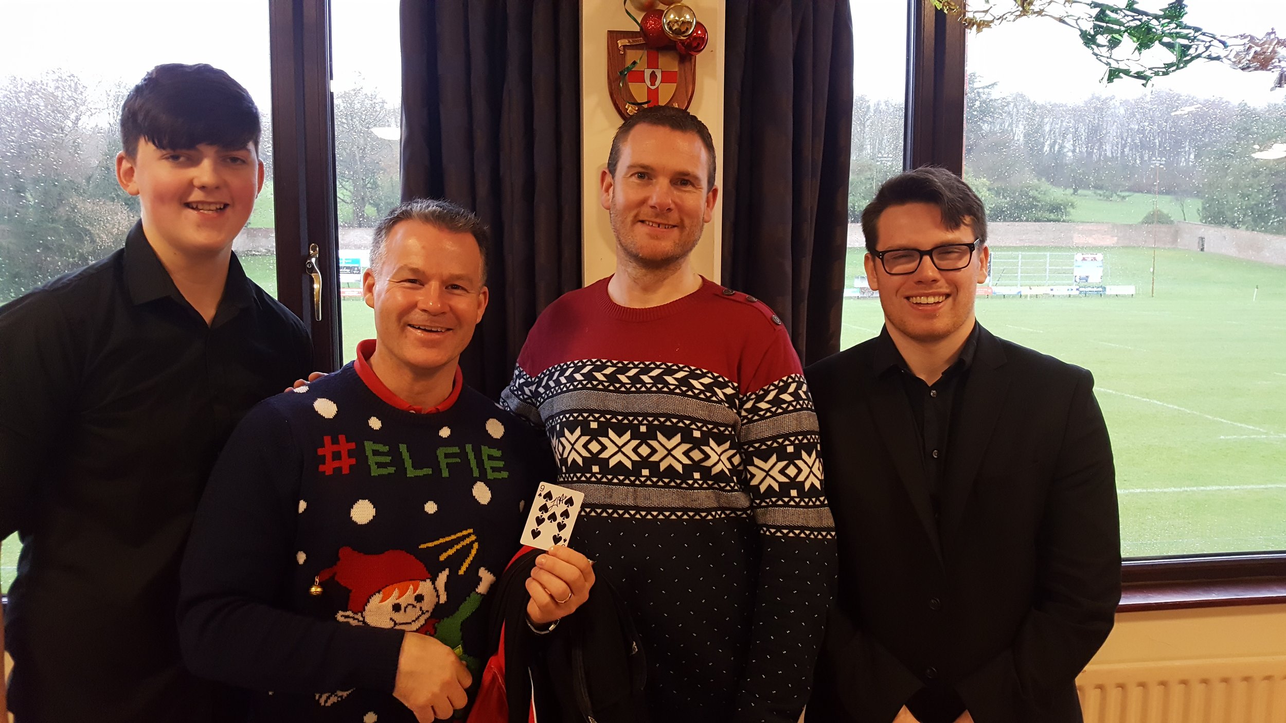 Four men standing together indoors, in front of a window overlooking a rainy outdoor sports field. One man is wearing a festive Christmas sweater, while others are in dark clothing; one man is holding a playing card.