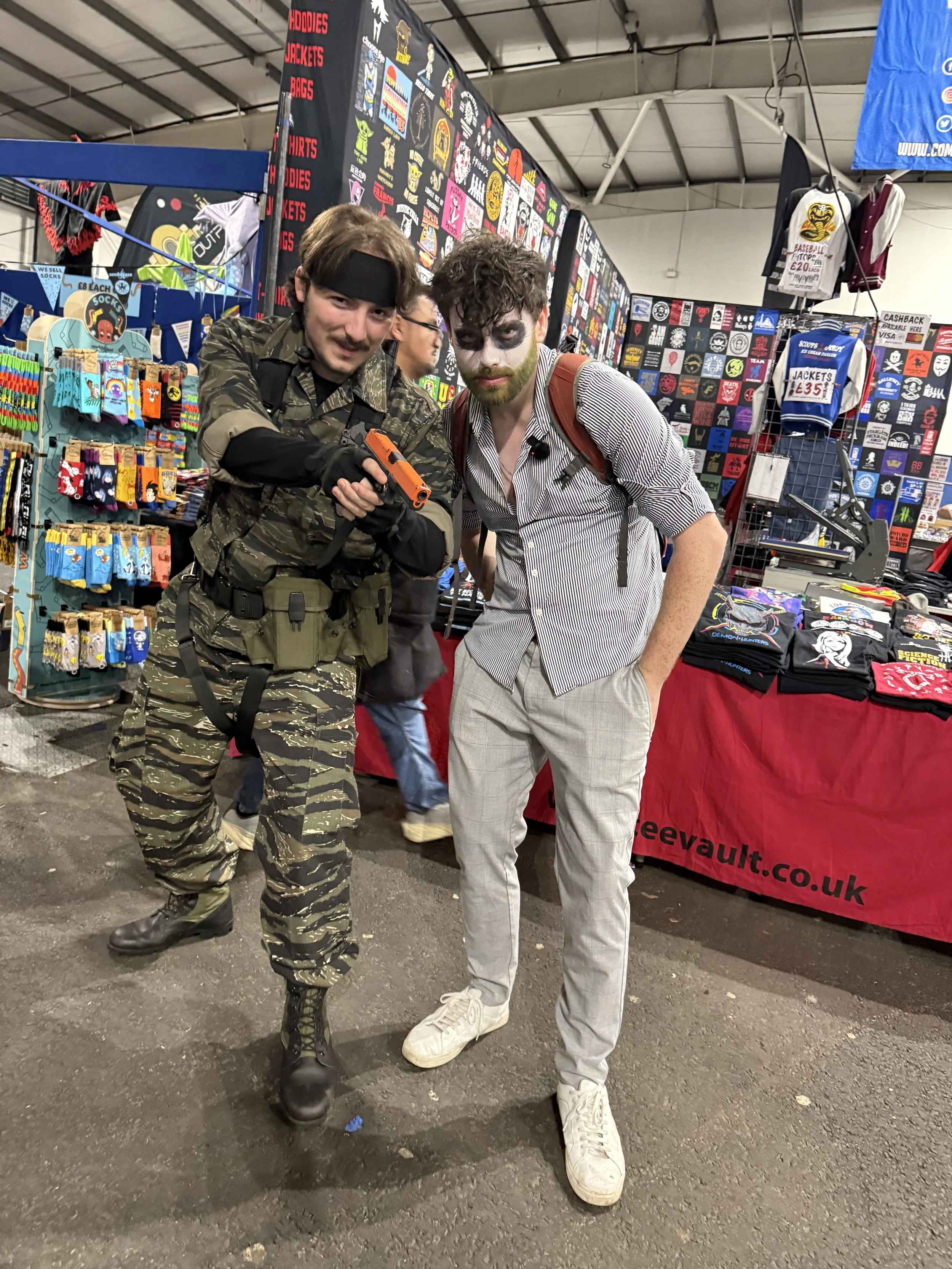 Two men in costume, one dressed as a soldier with camouflage clothing and a toy gun, the other with face paint and casual clothes, standing at a booth at a convention or market, with colorful merchandise and posters in the background.